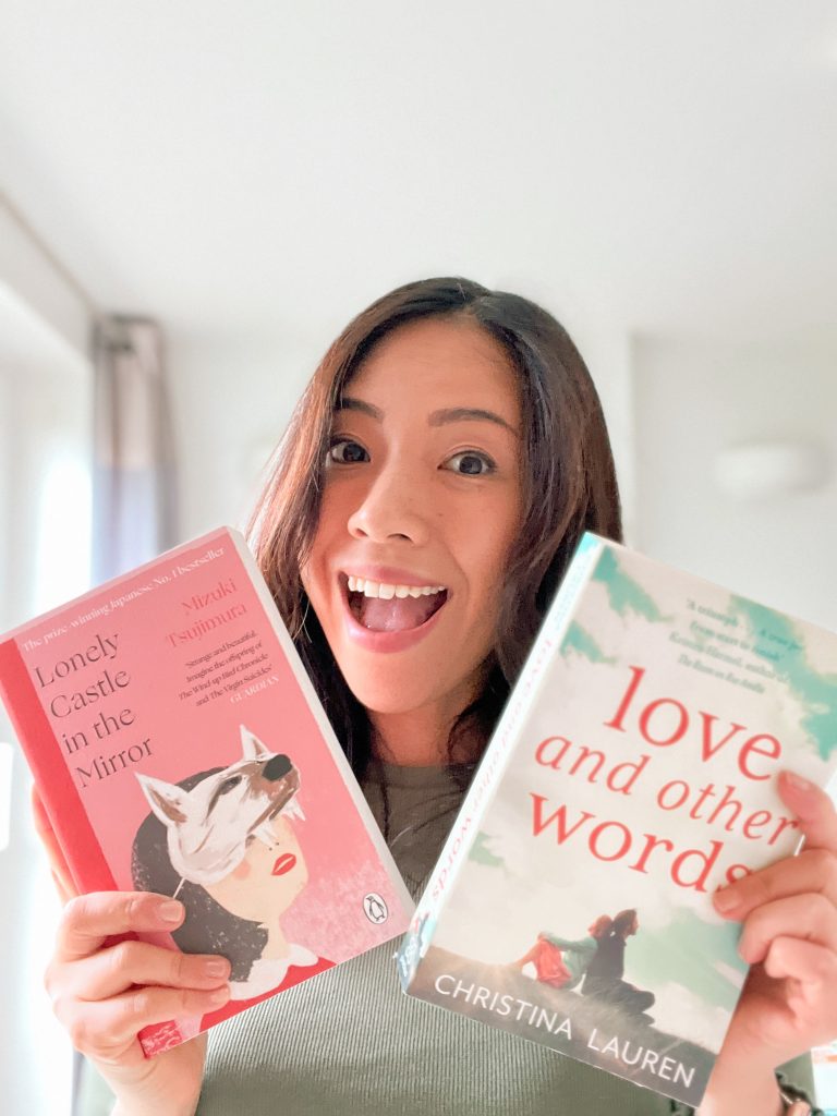 Smiling photo of Mitsy holding two books. In one hand, she is holding a copy of the book Lonely Castle in the Mirror by Mizuki Tsujimura, a pink bok cover with a woman wearing a white wold mask. In her other hand, she is holding a copy of the book Love and Other Words by Christina Lauren, a white book cover with parts of a blue sky.