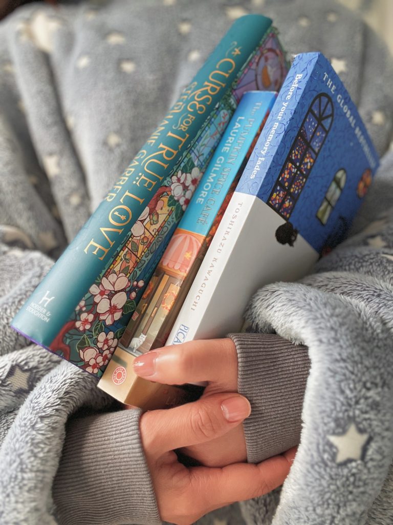 A close up image of Mitsy's hands cradling four books in her arms. She is wearing a soft gray sweater with white stars on it. The books are various shades of blue to match.