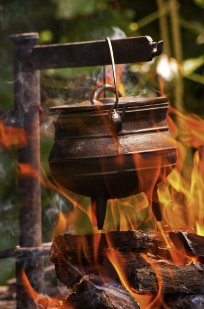 An iron pot hovers over an open flame. The pot and fire are seemingly outdoors, and the flame engulfs the pot.