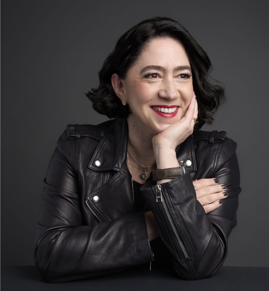 Caroline Topperman, a dark haired, light skinned woman clad in a leather jacket smiles to the side of the camera while propping her cheek and chin up with her hand.