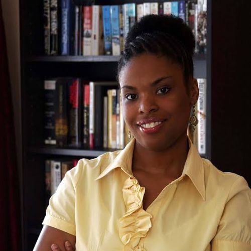 A dark brown skinned woman smiles into the camera wearing a yellow ruffled polo in front of a black book case filled with paperbacks.