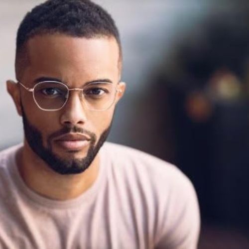 A light skinned black man in glasses and a trimmed beard, wearing a tan t-shirt looks at the camera.