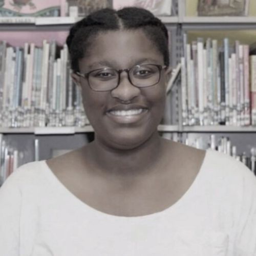 A Dark brown skinned woman waring rectangle glasses and a scoop necked shirt smiles at the camera before shelves of children's books