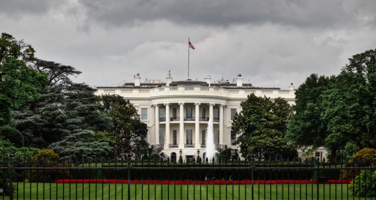 The White House in Washington, DC, as viewed through the fence across the back lawn; cloudy skies are overhead.