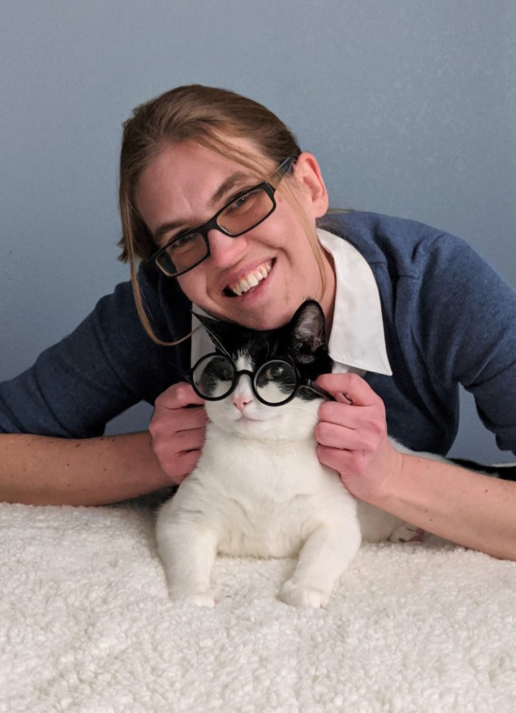 Portrait of Leslie Vedder. She is wearing a blue shirt and wearing glasses. Her cat below her is also wearing glasses, looking at the camera.