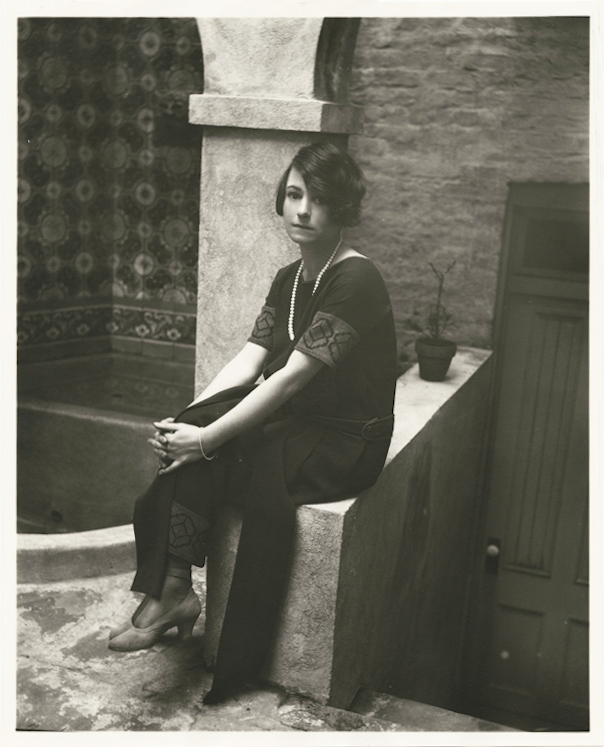 In black and white, Dorothy Parker wears a short-sleeved black dress with embroidered detail, a pearl necklace, and high heels. She sits perched on a low wall. A building with a door, column, potted plant, and hung tapestry are in the background.