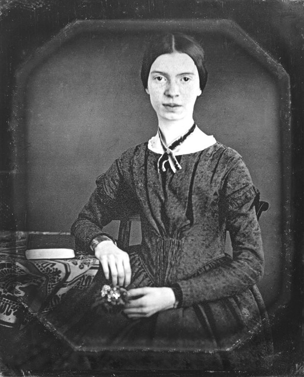 In black and white, Emily Dickinson sits at a table with a printed tablecloth and a book. She wears a black dress with a black ribbon around her neck and holds a flower in her hand.