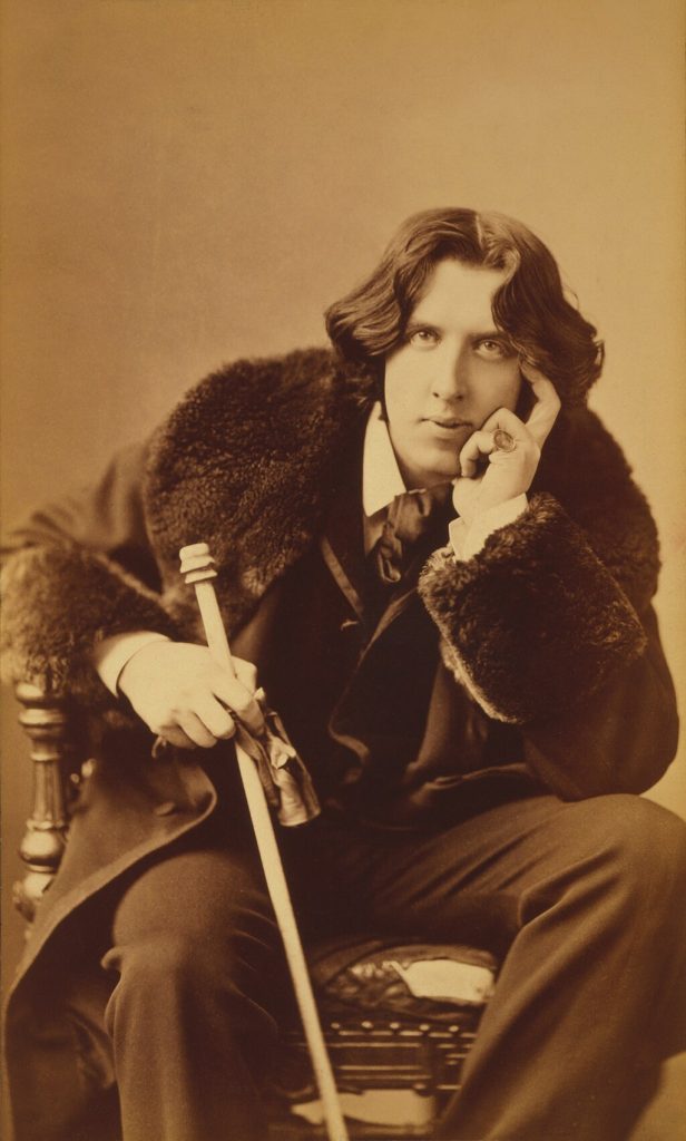 Oscar Wilde sits in a chair in a suit and fur-trimmed coat. He holds a walking stick and rests his chin on his hand. A large signet ring is on his pinkie finger.