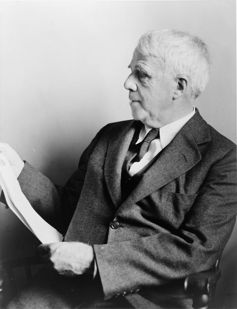 In black and white, Robert Frost sits and reads a paper in his hand. He wears a suit.