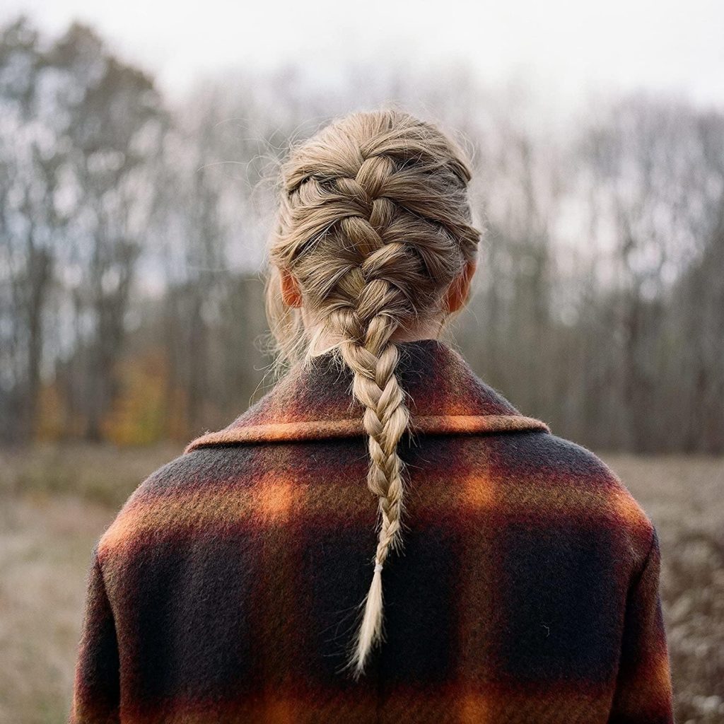 The album cover for evermore by Taylor Swift. Taylor Swift is depicted from behind, wearing an orange and dark navy coat with her blonde hair in a French braid. She stands in a forest clearing in late fall, with most of the trees missing leaves.