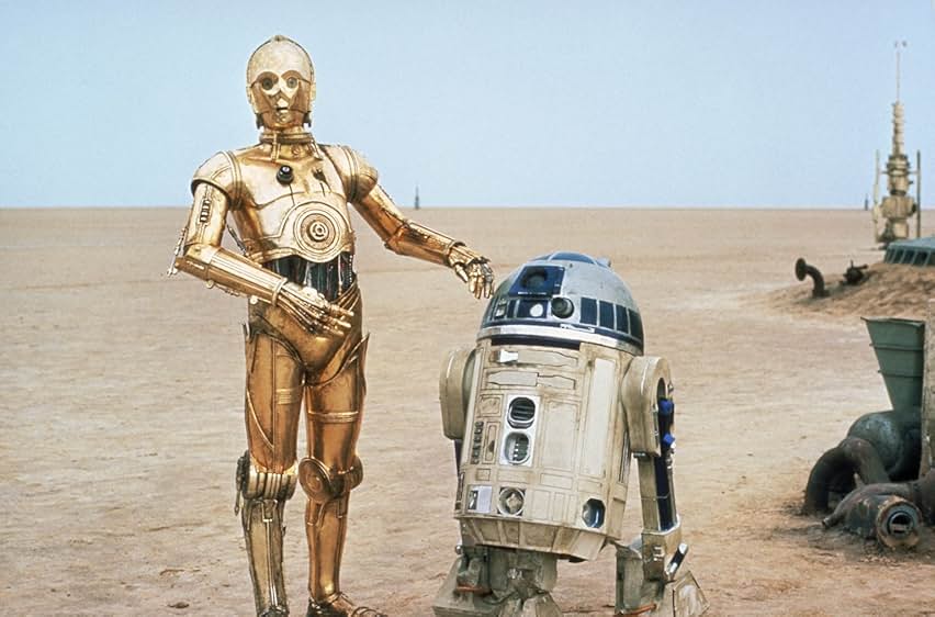 A desert landscape with a pale blue sky. Some machinery in the background to the right. C-3PO and R2-D2 stand beside each other.