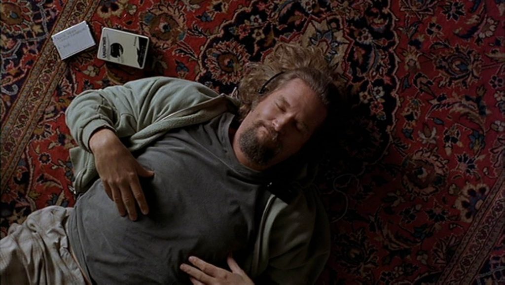 A red rug with black and yellow decorative designs. A man lies on his back with his eyes closed and black headphones on. He wears a gray t-shirt, gray shorts, and a green hoodie. A cassette player and cassette tape are beside him.