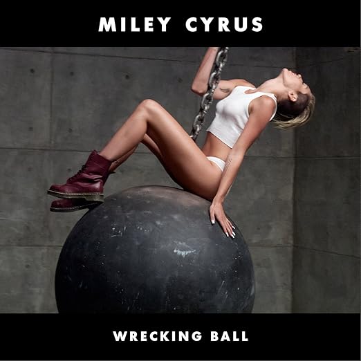 Miley Cyrus wears a white cropped tank top, white underwear, and red leather boots. She sits on top of a wrecking ball in front of a cement wall. Wrecking Ball in white lettering.