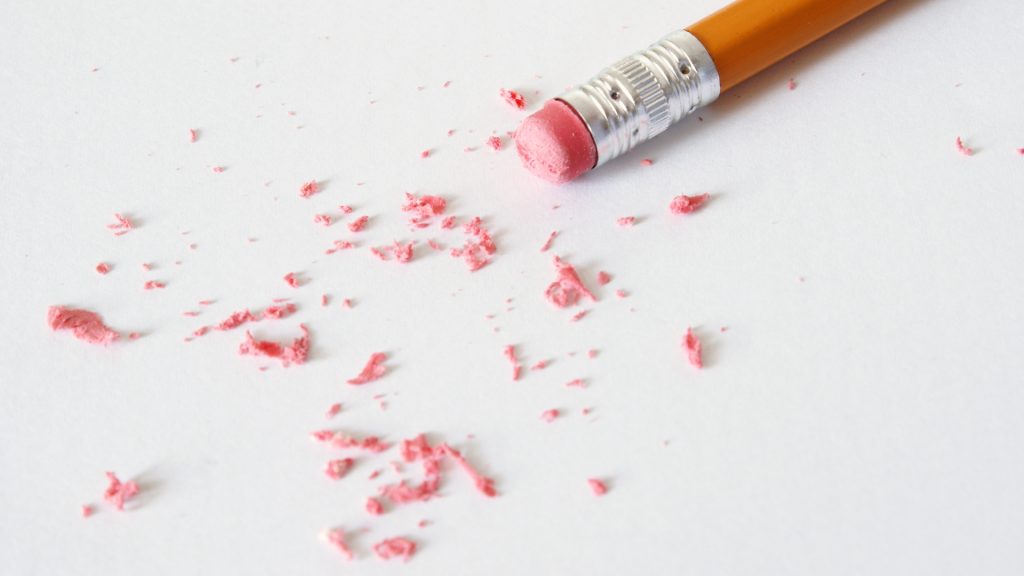 A white surface. A yellow pencil with a worn down pink eraser. Pink eraser shavings are scattered across the surface.