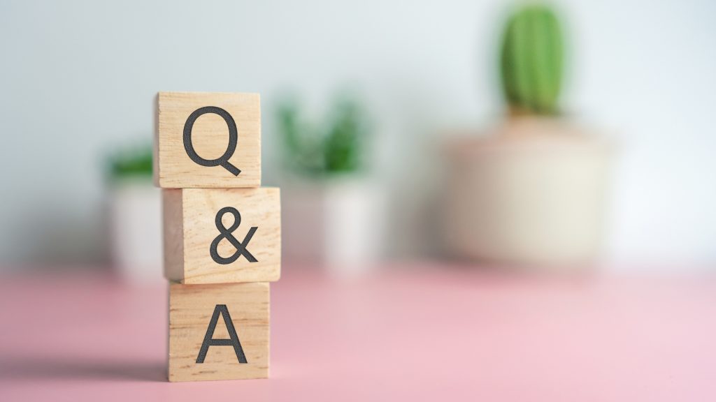 Three wooden cubes stacked on each other with Q, &, and A carved into them. A blurred background with a pink table and three potted cacti.