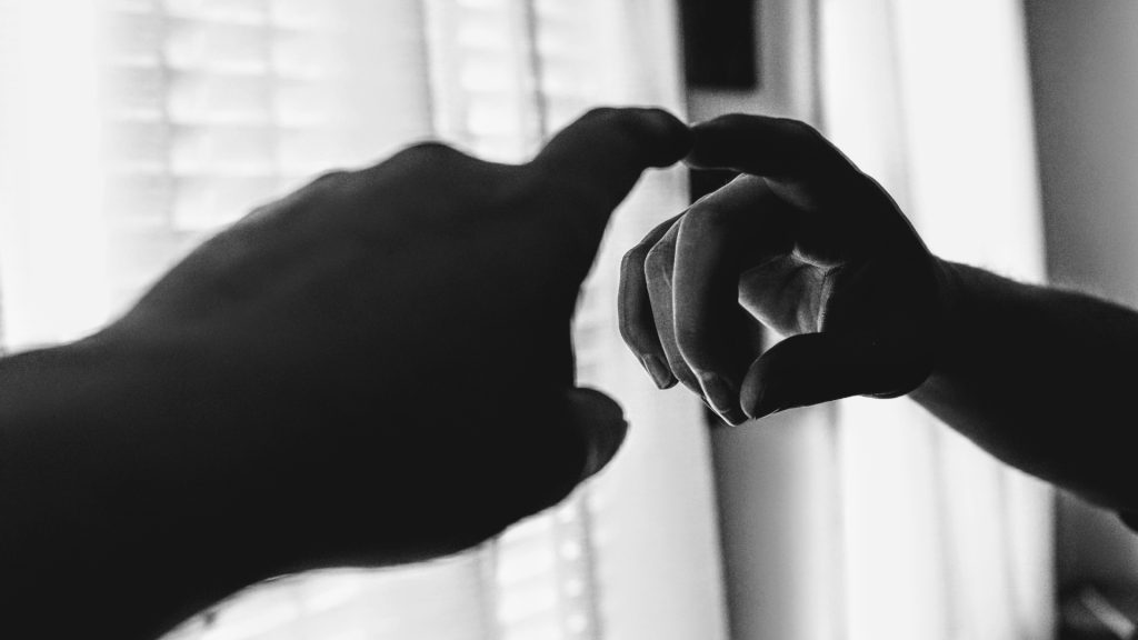 In black and white, a hand reaches forward and touches its reflection in a mirror. A curtained window in the background.
