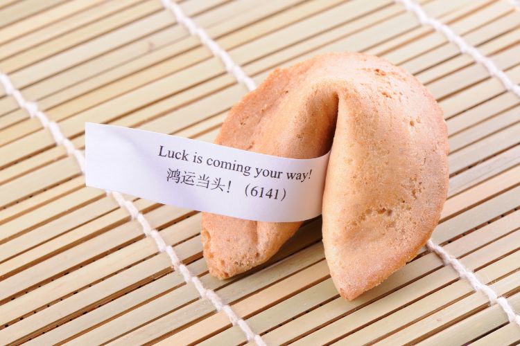 A fortune cookie with the fortune slip that says "Luck is coming your way! (6141)" and some Chinese characters. The cookie is sitting on a bamboo mat.