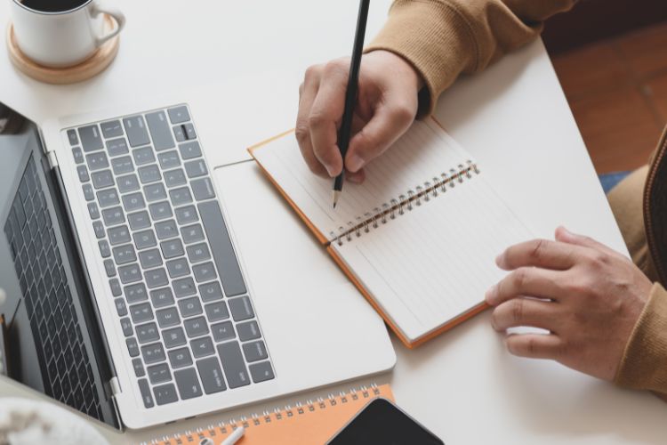 Someone writing in a small notebook in front of a laptop.
