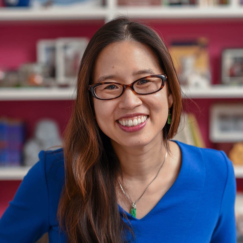 Grace Lin, a smiling tan-skinned woman with long, straight auburn hair, wearing glasses and a blue blouse, standing in front of a red wall with white bookshelves.