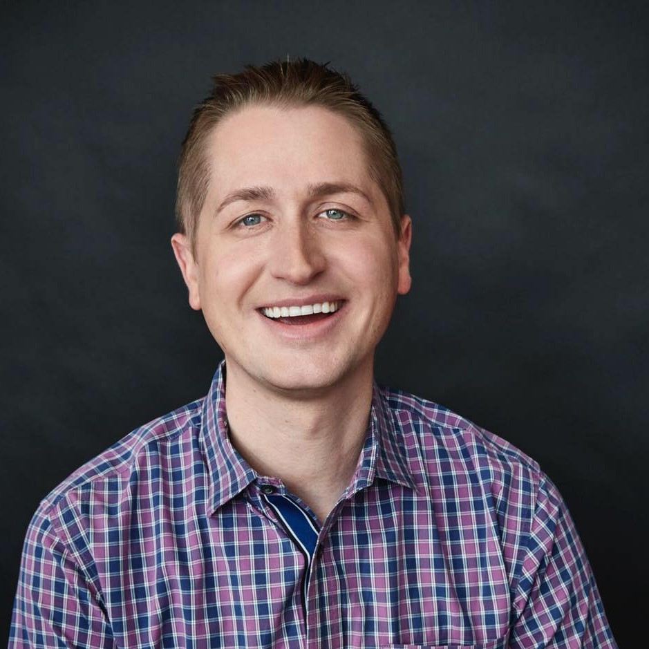John Schu, a smiling light-skinned man with short blond hair and blue eyes wearing a blue and purple checked button-down shirt sitting in front of gray background.