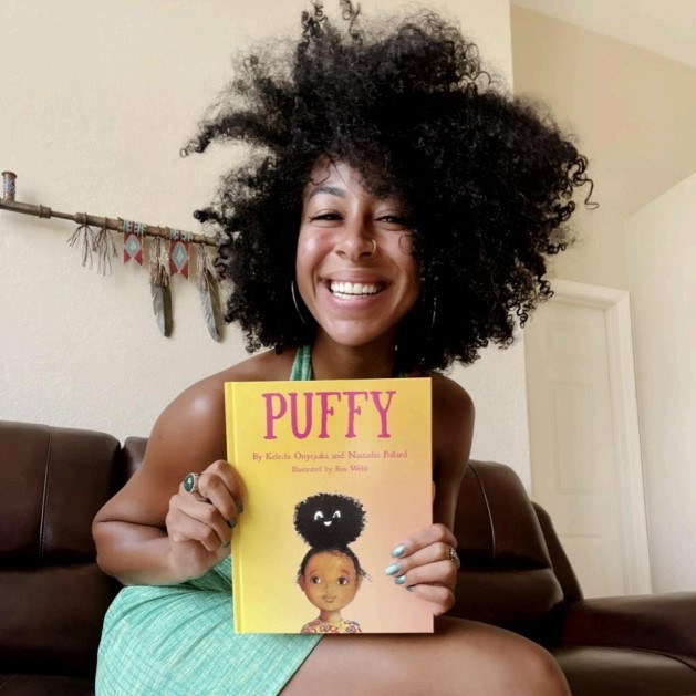 Nastasha Pollard, a smiling brown-skinned woman with dark, coily hair in an afro style, sitting on a brown leather couch and displaying her book Puffy on her lap.