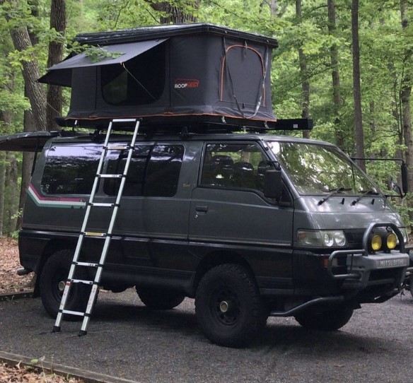 A black 90s-era Mitsubishi camper van with pop-up sleeping tent on its roof parked in a wooded area.