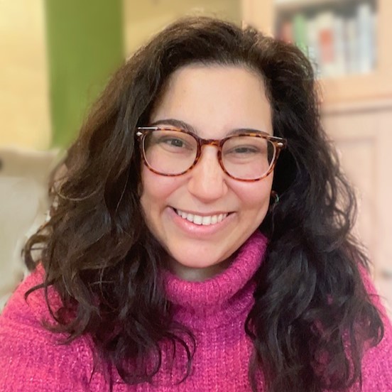 Tyler Feder, a smiling light-skinned woman with long, wavy dark hair, wearing eyeglasses and a pink sweater in front of a green wall and some bookshelves.
