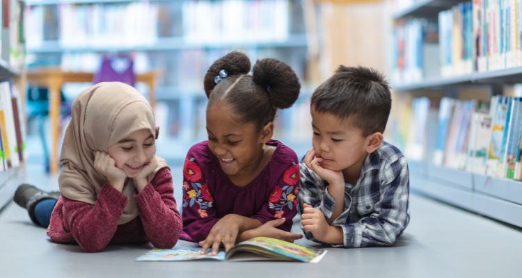Three smiling children diverse in skin tone and hair color/texture — and in hijab — lay prone on the floor of a library and read a book together.