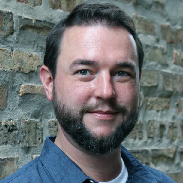 Alex Willan, a smiling light-skinned adult man with short brown hair and beard, wearing a blue collared shirt white standing next to a brick wall.