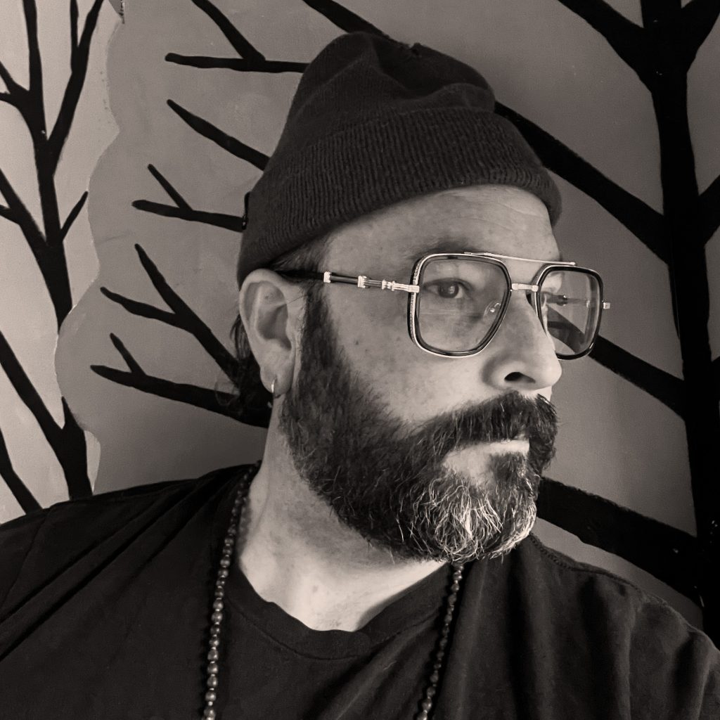 Black-and-white photo of Zachariah OHora, a light-skinned man with dark beard and mustache wearing glasses and a knit hat, looking off to the right while sitting in front of a wall decorated with stencils of tree limbs.