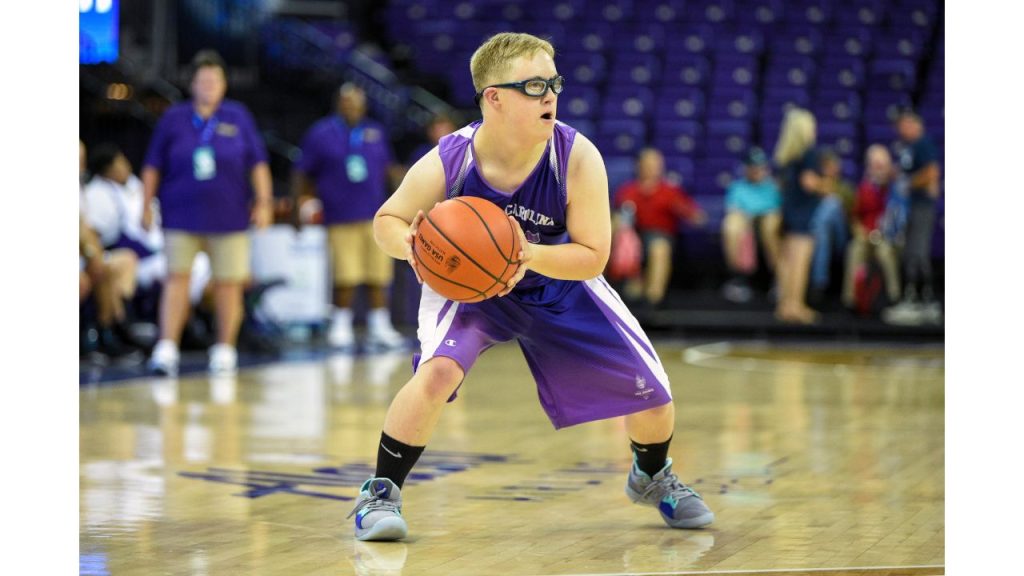 A young man with down syndrome is holding a basketball in a purple uniform.