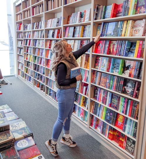 lottie-image-6 Bookstagrammer Lottie with a book in her arms as she reaches up while on her tip toes to look at a book on a bookshelf covering the wall of the store.
