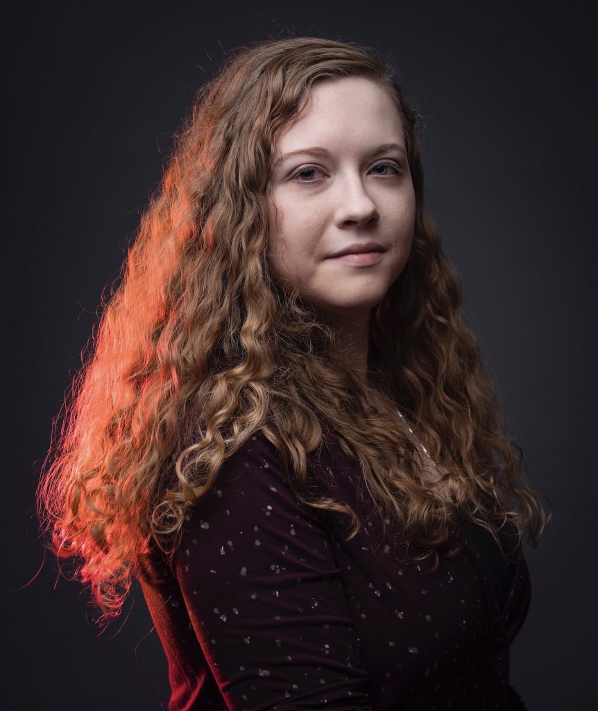 A black background. Lynn Buchanan wears a burgundy top with with a small gray dotted pattern. She is illuminated by a red light coming from the left.
