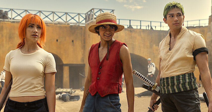 Live-action scene from Netflix's 'One Piece' series, showcasing main characters Nami, Luffy, and Zoro standing together in a battle-worn environment, dressed in their iconic outfits.