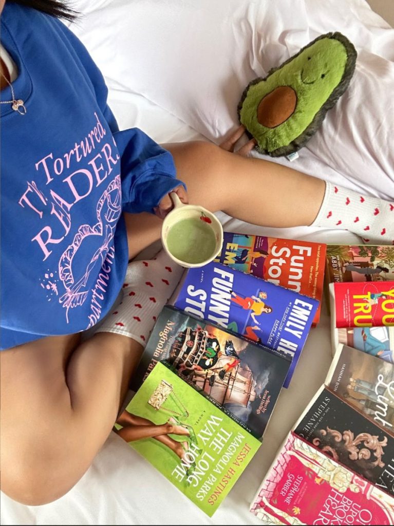 A picture of Joceline's lap holding a cup of mocha tea and books spread in front of her on her bed. An avocado jellycat is laying next to her. Joceline is wearing a blue bookish sweater and white socks with hearts on them.