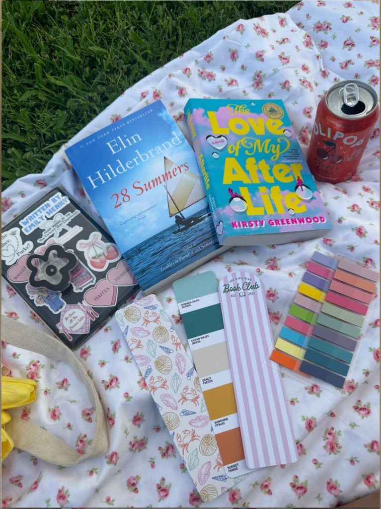 A picture of books layed out on a blanket in the grass, an olipop can is next to them, along with bookmarks and sticky pads.