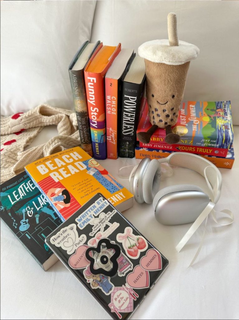 A picture of books stacked and placed on Joceline's bed. A boba drink jelly cat is sitting on one of the stacks, and white headphones with a ribbon bow tied on them is set next to the books.