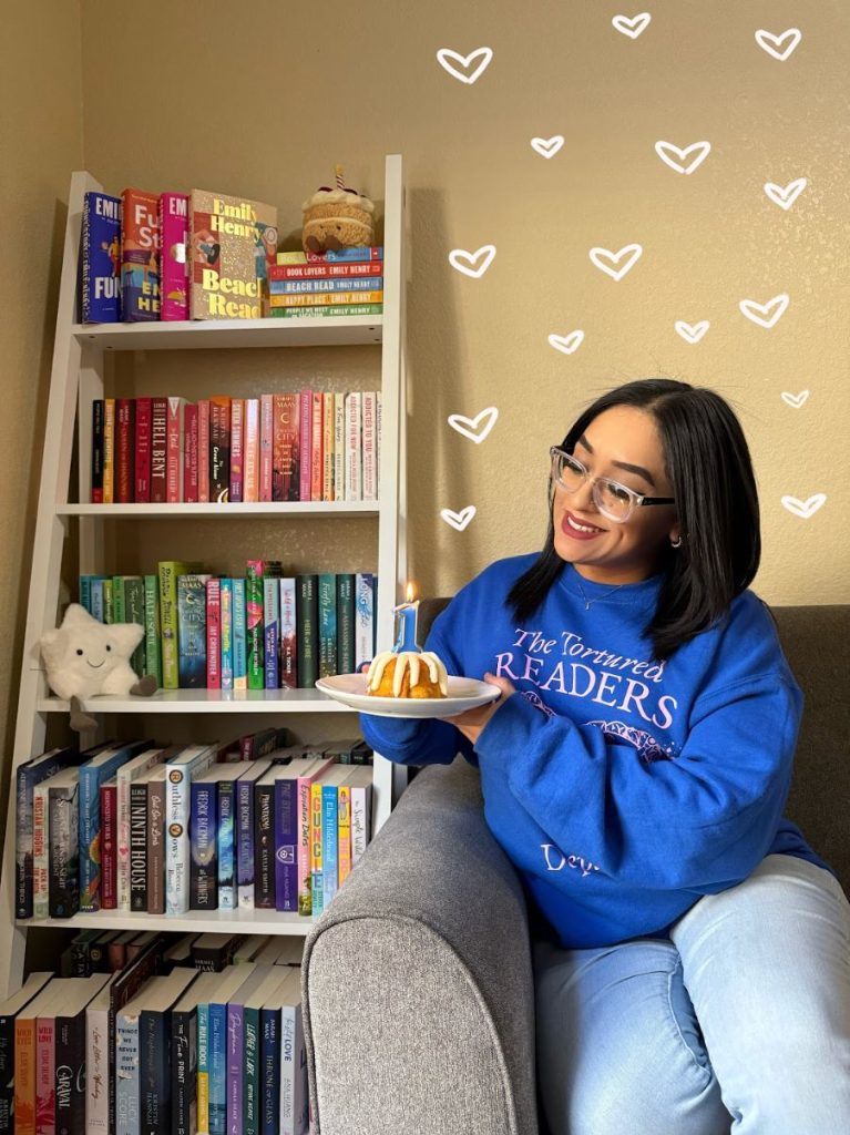 Picture of Joceline @bookishlyjoceline sitting on a couch next to a bookshelf and holding a small cake with the a number "1" candle. White drawn hearts are placed all above her.