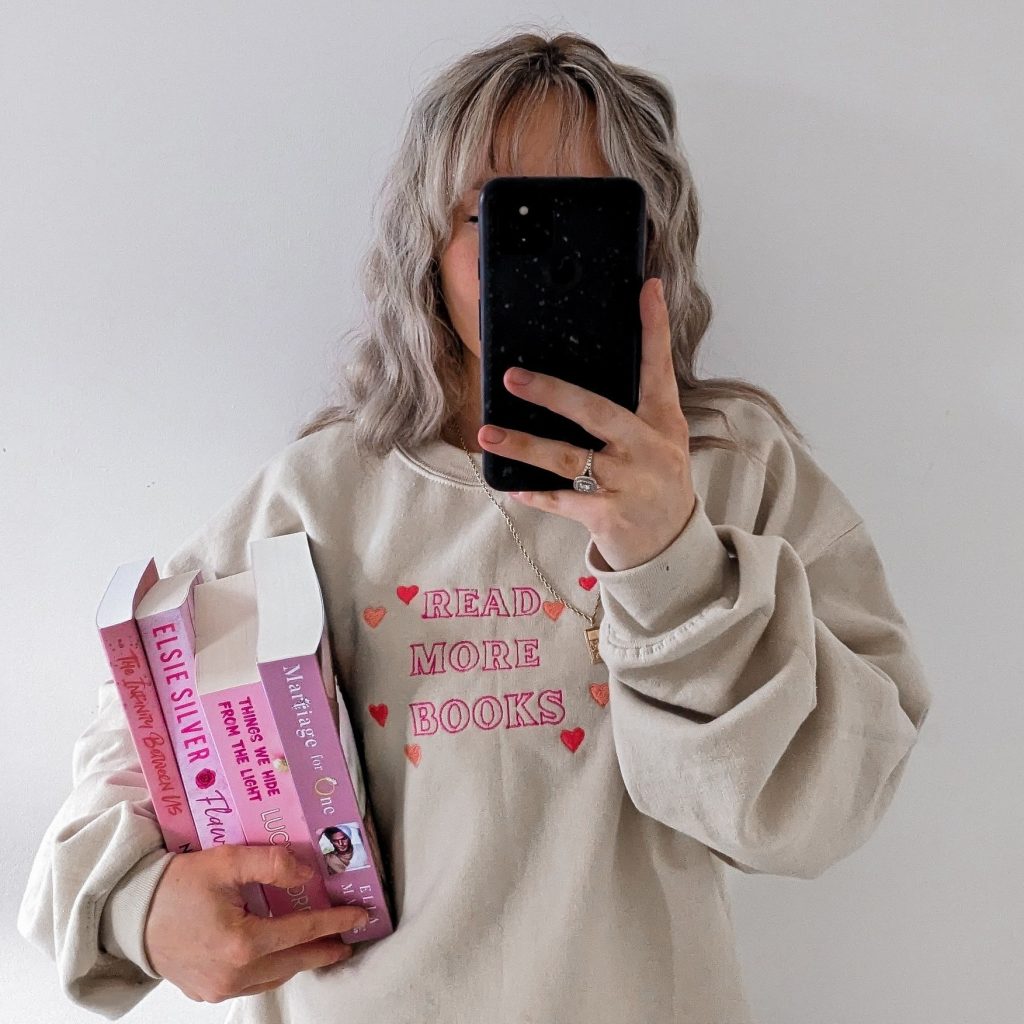 lottie-photo-one Bookstagrammer Lottie taking a mirror selfie with her black phone covering her face. In her arms is a stack of four pink books complementing the pink words on her tan sweatshirt that says “Read More Books” with hearts around it.