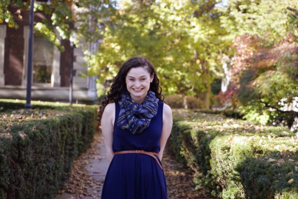 A park full of trees and low cut hedges. A path cutting through the hedges is littered with fallen leaves. In the back, there's a part of a white stone building. Christine Calella smiles and wears a blue dress with a skinny brown leather belt and a blue and green striped scarf.