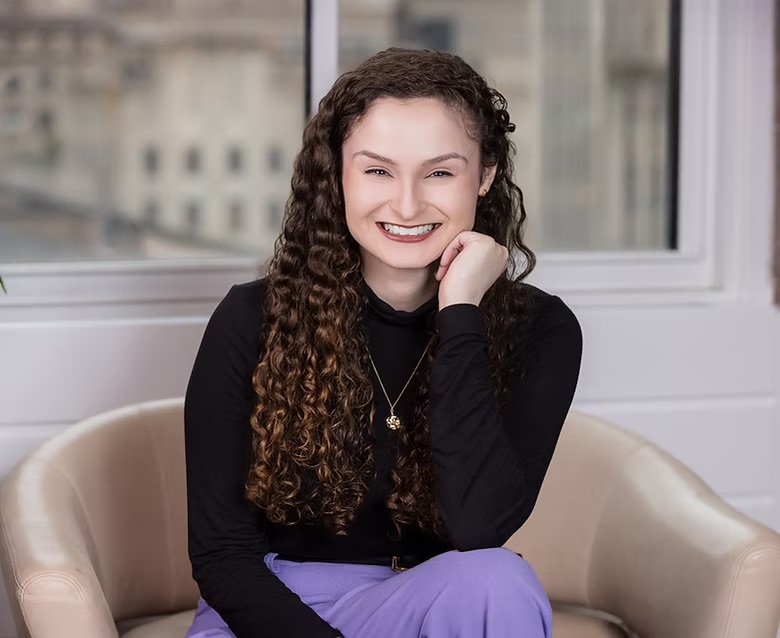 A white wall with a window looks out onto a blurred background. Christine Calella sits in a beige leather chair and wears a black shirt, purple skirt, and gold necklace. She smiles and rests her chin on her hand.