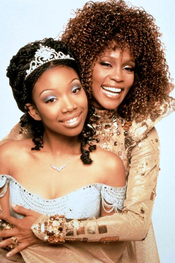 The background is pale blue. Brandy smiles and wears a crown, necklace, and a blue sparkly ballgown. Whitney Houston smiles and hugs her from behind. She wears a gold glittery gown.