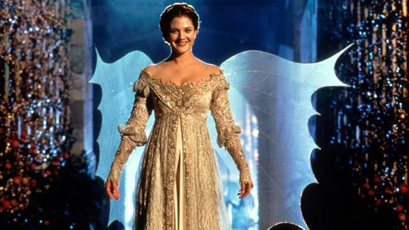 Two walls covered with flowers and glittering decorations. In the middle stands a smiling woman in a white jeweled gown with transparent fairy wings.