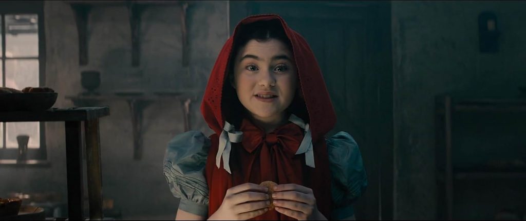 The inside of a cottage with a wood door and stone walls. A young girl with dark hair tied in blue ribbons wears a blue dress and red hooded cape. She holds a cookie in her hands.