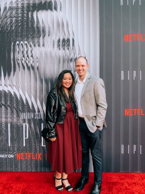 Natalie and her husband Jared standing on a red carpet in front of the grayscale movie poster for Netflix's Ripley. Natalie is wearing a red dress and leather jacket. Her husband, Jared, is wearing a gray coat and black pants.