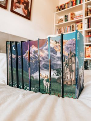 The Throne of Glass special edition box set sits on a bed with white covers. The box set consists of eight books lined side by side. On the edges of their pages, a mountain landscape is painted with a sky progressing from night to day, left to right. The landscape includes a white deer, flying eagle, tree, and castle.
