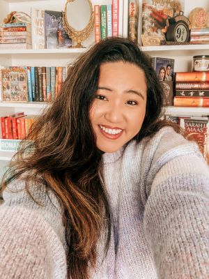A selfie of Bookstagrammer Natalie, an Asian woman, standing in front of a white bookshelf. Natalie smiles at the viewer, holding the camera with both hands. She has long dark hair swept over one shoulder and a gray sweater.
