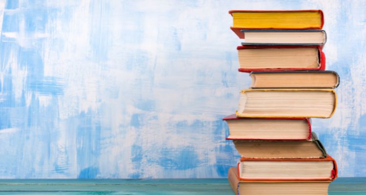 A stack of books sits on a blue surface. Only the edges of the pages are visible. The background is painted in blue and white splotches.