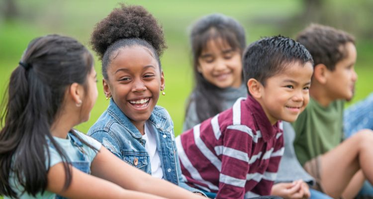 A smiling group of children diverse in skin tone, hail color/texture, and facial features, sitting outside in a green space.