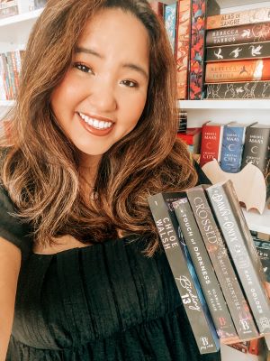 Selfie of Natalie smiling at the viewer while standing in front of a bookshelf. Natalie is holding four books: Binding 13 by Chloe Walsh, Touch of Darkness by Scarlett St. Clair, Crossed by Emily McIntire, and A Dawn of Onyx by Kate Golden.